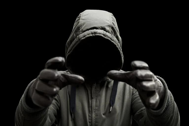 A terrifying mysterious man wearing a hooded jacket with arms raised in a strangling or attacking pose. Shot against a black background.